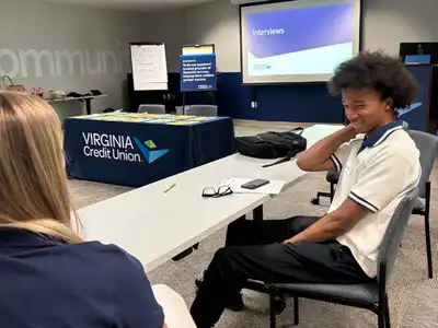 Blonde woman with back to camera sits opposite young Black man in white polo with the word 